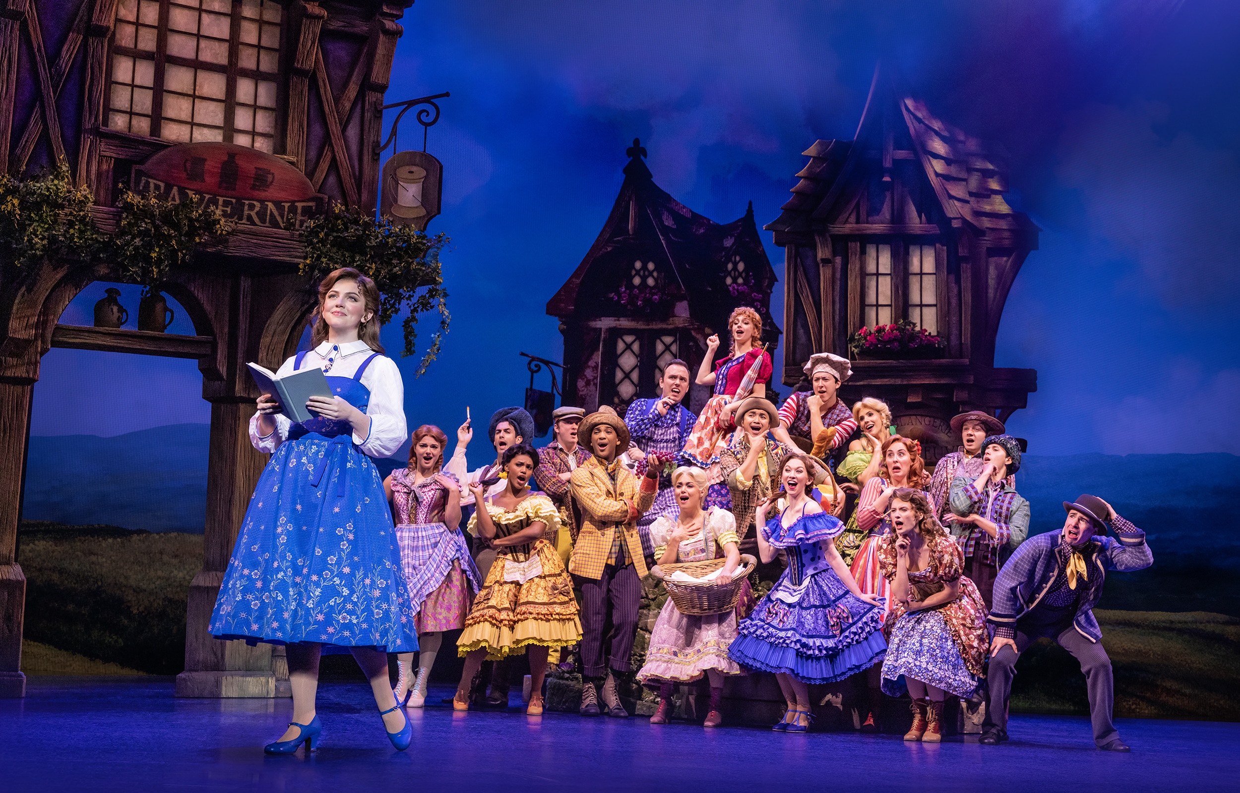 Belle reads a book as villagers gather behind her during the opening village scene in Disney's Beauty and the Beast.
