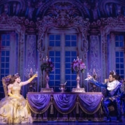Belle and the Beast share a candlelit castle scene in Disney's Beauty and the Beast, seated across an elegant dining table in a lavish ballroom setting.
