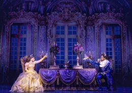 Belle and the Beast share a candlelit castle scene in Disney's Beauty and the Beast, seated across an elegant dining table in a lavish ballroom setting.