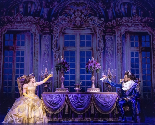Belle and the Beast share a candlelit castle scene in Disney's Beauty and the Beast, seated across an elegant dining table in a lavish ballroom setting.