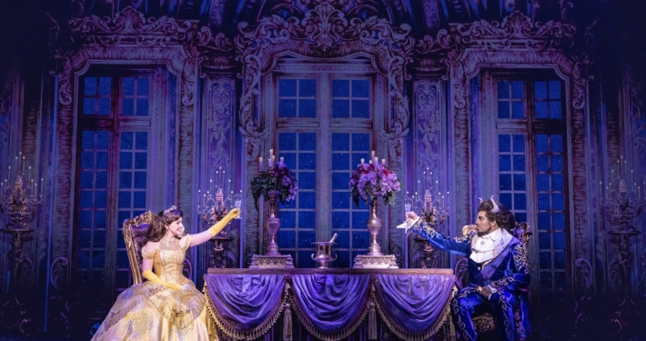 Belle and the Beast share a candlelit castle scene in Disney's Beauty and the Beast, seated across an elegant dining table in a lavish ballroom setting.