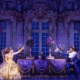 Belle and the Beast share a candlelit castle scene in Disney's Beauty and the Beast, seated across an elegant dining table in a lavish ballroom setting.
