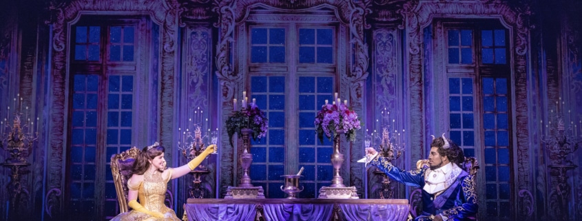 Belle and the Beast share a candlelit castle scene in Disney's Beauty and the Beast, seated across an elegant dining table in a lavish ballroom setting.
