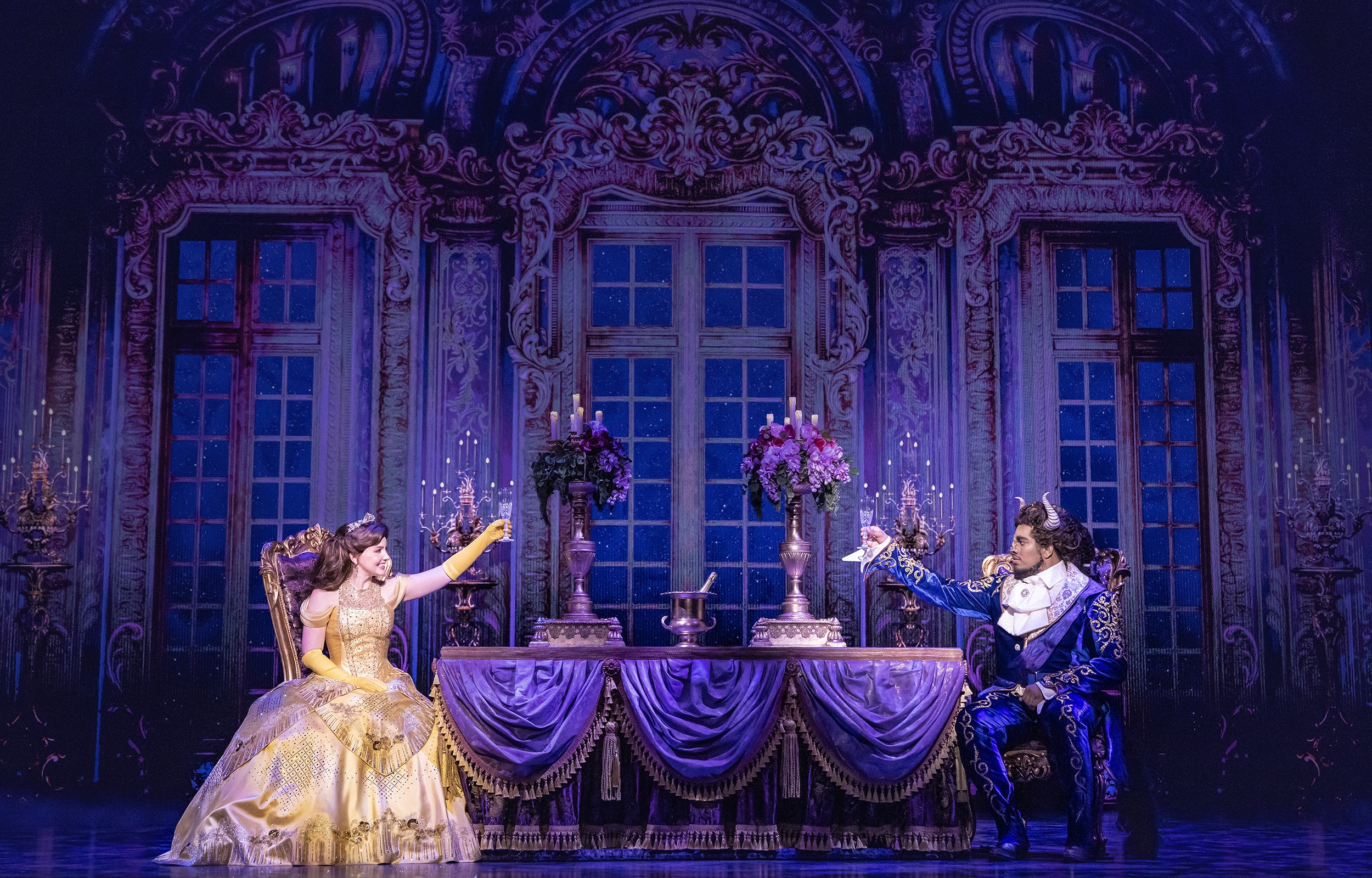 Belle and the Beast share a candlelit castle scene in Disney's Beauty and the Beast, seated across an elegant dining table in a lavish ballroom setting.