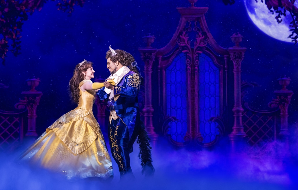 Belle and the Beast dance beneath a moonlit sky in Disney's Beauty and the Beast, surrounded by a romantic castle backdrop and theatrical fog.