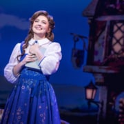 Belle stands center stage holding a book in Disney's Beauty and the Beast, wearing a blue village dress against a charming French village backdrop.