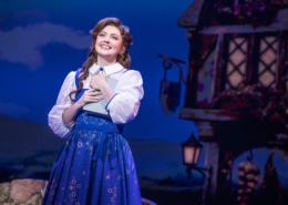 Belle stands center stage holding a book in Disney's Beauty and the Beast, wearing a blue village dress against a charming French village backdrop.