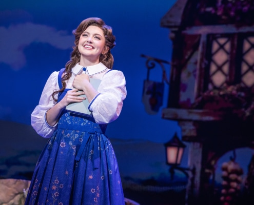 Belle stands center stage holding a book in Disney's Beauty and the Beast, wearing a blue village dress against a charming French village backdrop.