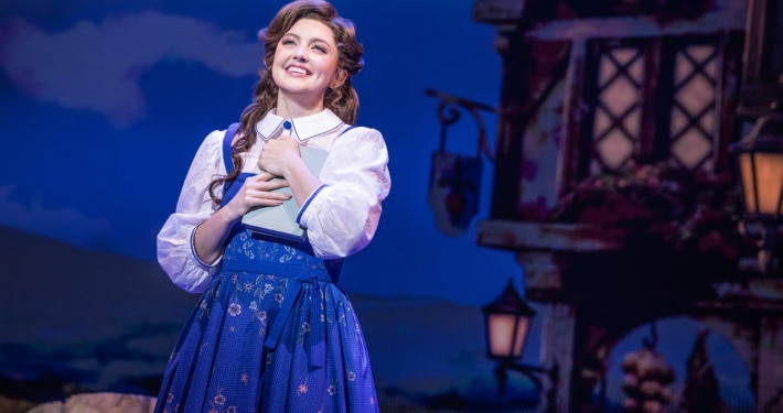 Belle stands center stage holding a book in Disney's Beauty and the Beast, wearing a blue village dress against a charming French village backdrop.