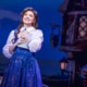 Belle stands center stage holding a book in Disney's Beauty and the Beast, wearing a blue village dress against a charming French village backdrop.