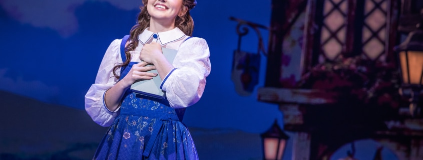 Belle stands center stage holding a book in Disney's Beauty and the Beast, wearing a blue village dress against a charming French village backdrop.