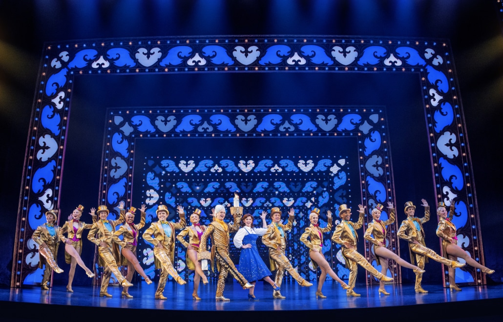 Disney's Beauty and the Beast national tour cast performs a lavish ensemble dance number on a blue illuminated stage with ornate set designs and gold costumes.