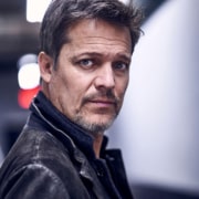 Professional headshot of a person wearing a black leather jacket over a dark shirt. The portrait is taken in an indoor setting with a softly blurred background and dramatic, directional lighting.