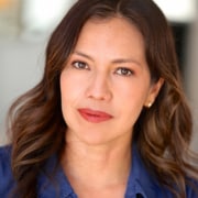 Professional headshot of a person with shoulder-length wavy dark hair wearing a blue collared shirt and small stud earrings. The portrait is photographed with natural lighting against a softly blurred background, creating a clean and professional appearance.