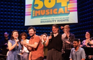 Group of performers and creators gathered onstage during a live event for 504 The Musical. Several participants hold microphones while others stand or sit nearby. A large screen behind them displays the show's title and the text “Inspired by the 1977 Sit-In for Disability Rights.”