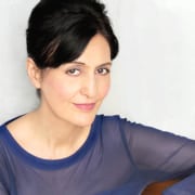 Professional headshot of a person with dark hair styled up, wearing a sheer blue long-sleeved top. The portrait is framed from the shoulders up against a light gray background, with the subject angled slightly toward the camera and illuminated by soft, even studio lighting.