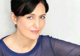 Professional headshot of a person with dark hair styled up, wearing a sheer blue long-sleeved top. The portrait is framed from the shoulders up against a light gray background, with the subject angled slightly toward the camera and illuminated by soft, even studio lighting.