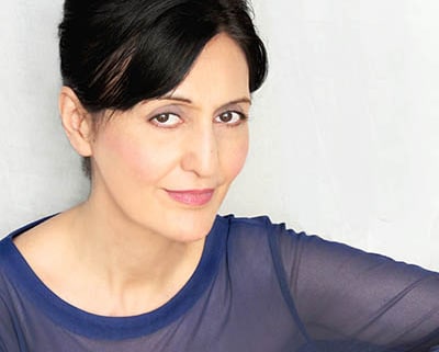 Professional headshot of a person with dark hair styled up, wearing a sheer blue long-sleeved top. The portrait is framed from the shoulders up against a light gray background, with the subject angled slightly toward the camera and illuminated by soft, even studio lighting.