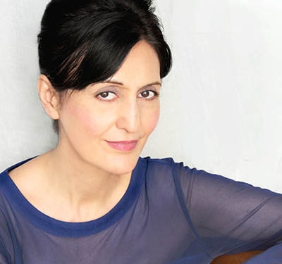 Professional headshot of a person with dark hair styled up, wearing a sheer blue long-sleeved top. The portrait is framed from the shoulders up against a light gray background, with the subject angled slightly toward the camera and illuminated by soft, even studio lighting.