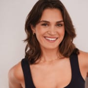 Professional headshot of a person with shoulder-length brown hair wearing a dark sleeveless top and a delicate necklace. The portrait is framed from the chest up against a light neutral background, with soft lighting and a clear focus on the face.