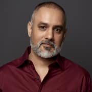 Professional headshot of a person with closely cropped hair and a gray beard, wearing a burgundy button-front shirt. The portrait is set against a dark gray studio background and framed from the chest up with soft, even lighting.