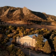 Aerial view of a theater building nestled among trees at the base of dramatic sandstone mountain formations during golden hour. The light-colored building sits in a wooded area with autumn foliage, while the rugged mountains rise prominently in the background under a clear blue sky. Warm evening sunlight casts long shadows across the landscape, highlighting the contrast between the forested valley and the rocky peaks.