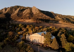 Aerial view of a theater building nestled among trees at the base of dramatic sandstone mountain formations during golden hour. The light-colored building sits in a wooded area with autumn foliage, while the rugged mountains rise prominently in the background under a clear blue sky. Warm evening sunlight casts long shadows across the landscape, highlighting the contrast between the forested valley and the rocky peaks.