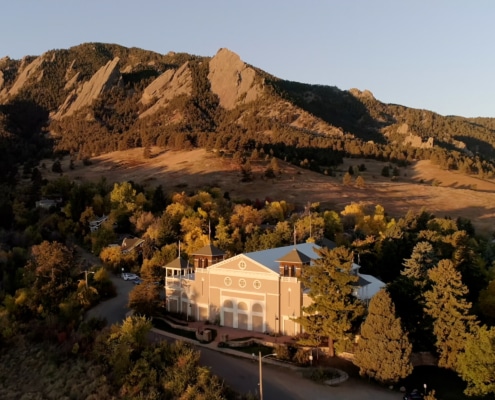 Aerial view of a theater building nestled among trees at the base of dramatic sandstone mountain formations during golden hour. The light-colored building sits in a wooded area with autumn foliage, while the rugged mountains rise prominently in the background under a clear blue sky. Warm evening sunlight casts long shadows across the landscape, highlighting the contrast between the forested valley and the rocky peaks.