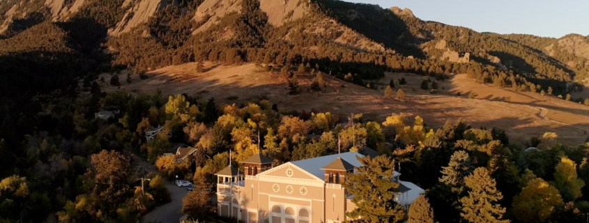 Aerial view of a theater building nestled among trees at the base of dramatic sandstone mountain formations during golden hour. The light-colored building sits in a wooded area with autumn foliage, while the rugged mountains rise prominently in the background under a clear blue sky. Warm evening sunlight casts long shadows across the landscape, highlighting the contrast between the forested valley and the rocky peaks.