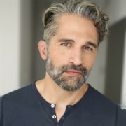 Professional headshot of a person with salt-and-pepper hair and a neatly trimmed beard, wearing a dark henley-style shirt. The portrait is framed from the shoulders up against a softly blurred neutral background, with even lighting highlighting facial features.