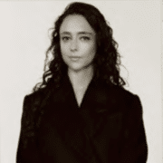 Black-and-white portrait photograph of a person with long, curly hair wearing a dark jacket, centered against a plain light background. The portrait appears printed on a vintage-style, cream-colored photo card with scalloped edges, displayed against a blue textured backdrop.