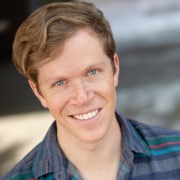 Professional headshot of a person with light-colored hair wearing a striped button-down shirt. The portrait is framed from the shoulders up against a softly blurred background with natural lighting.