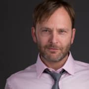 Professional headshot of a person with short brown hair and a neatly trimmed beard, wearing a light pink dress shirt and gray tie. The portrait is framed from the chest up against a dark gray background with studio lighting.