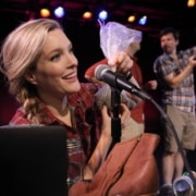 Stage production photo showing a performer seated at a table covered with props and sound-effect materials, speaking into a microphone while holding a small plastic bag near the microphone. Items in the foreground include a glass jar, a leather boot, and audio equipment used for live Foley effects. Two additional performers stand in the background on a dimly lit stage. Colored stage lights in red, blue, and purple illuminate the performance space.