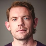 Professional headshot of a person with short light-colored hair wearing a dark crew-neck shirt. The portrait is framed from the shoulders up against a neutral gray background, with soft studio lighting and direct eye contact with the camera.