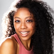 Professional headshot of a person with long curly hair wearing a sleeveless rose-colored top. The portrait is closely cropped against a light background, with bright studio lighting highlighting facial features and hair texture.