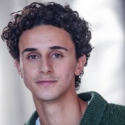 rofessional headshot of a person with short curly hair wearing a textured green sweater. The portrait is framed from the shoulders up against a softly blurred light background, with the subject facing the camera and illuminated by natural-looking light.
