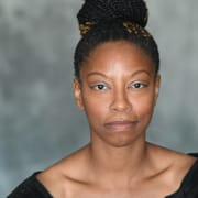 Professional headshot of a person with braided hair styled in a bun, wearing a black top. The portrait is framed from the shoulders up against a softly textured gray background with even studio lighting.