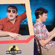 Stage production photo from Hold On To Your Butts showing two performers seated within a cardboard prop shaped like a vehicle. One performer, wearing sunglasses and a red checkered shirt, leans out of a window frame attached to the prop, while the other performer, wearing a plaid shirt, sits beside them reading from a sheet of paper. The vehicle prop features the show's logo on the side. Blue stage lighting illuminates the dark theater background.