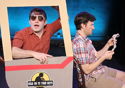 Stage production photo from Hold On To Your Butts showing two performers seated within a cardboard prop shaped like a vehicle. One performer, wearing sunglasses and a red checkered shirt, leans out of a window frame attached to the prop, while the other performer, wearing a plaid shirt, sits beside them reading from a sheet of paper. The vehicle prop features the show's logo on the side. Blue stage lighting illuminates the dark theater background.