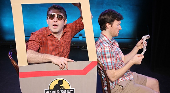 Stage production photo from Hold On To Your Butts showing two performers seated within a cardboard prop shaped like a vehicle. One performer, wearing sunglasses and a red checkered shirt, leans out of a window frame attached to the prop, while the other performer, wearing a plaid shirt, sits beside them reading from a sheet of paper. The vehicle prop features the show's logo on the side. Blue stage lighting illuminates the dark theater background.