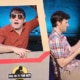 Stage production photo from Hold On To Your Butts showing two performers seated within a cardboard prop shaped like a vehicle. One performer, wearing sunglasses and a red checkered shirt, leans out of a window frame attached to the prop, while the other performer, wearing a plaid shirt, sits beside them reading from a sheet of paper. The vehicle prop features the show's logo on the side. Blue stage lighting illuminates the dark theater background.