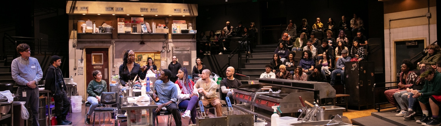 JamieKraus23_DCPA-ClydesStudentMatinee_140 Wide view of a theater-in-the-round performance space designed as a commercial kitchen. Audience members are seated on tiered risers surrounding the set, while a group of performers and participants gather around stainless-steel worktables and cooking equipment at center stage. Industrial lighting hangs overhead, illuminating the detailed kitchen environment with ovens, sinks, shelving, boxes, and supplies visible throughout the scene.