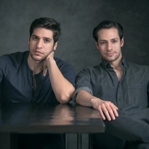 Studio portrait of two people seated at a dark table against a muted gray background. Both wear casual button-front shirts and face the camera. One rests their head on a hand while leaning an elbow on the table, and the other sits with an arm extended across the tabletop. Soft, even lighting highlights the subjects against the simple backdrop.