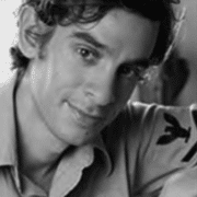 Black-and-white close-up portrait of a person with dark wavy hair wearing a light-colored collared shirt, looking toward the camera. The image is tightly cropped around the face and shoulders, with a softly blurred background.
