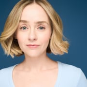 Professional headshot of a person with short, wavy blond hair wearing a light blue top. The portrait is framed from the shoulders up against a solid blue background with soft, even studio lighting.