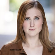 Professional headshot of a person with shoulder-length auburn hair wearing a brown jacket. The portrait is framed from the shoulders up against a softly blurred background with natural-looking light.
