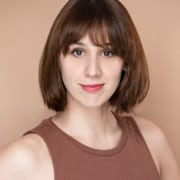 Professional headshot of a person with chin-length brown hair wearing a sleeveless brown top. The portrait is framed from the shoulders up against a warm beige background, with soft studio lighting and direct eye contact with the camera.
