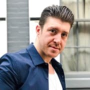 Professional headshot of a person with dark hair wearing a blue button-down shirt over a light-colored T-shirt. The portrait is photographed outdoors against a softly blurred urban background.