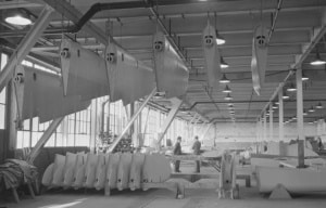 Black-and-white photograph of an aircraft manufacturing facility during wartime production. Multiple aircraft components, including fuselage and tail sections, hang from overhead conveyors while additional parts are stacked along the factory floor. Two workers stand near an assembly table in the center of the large industrial space, which is illuminated by rows of overhead lights and tall windows.